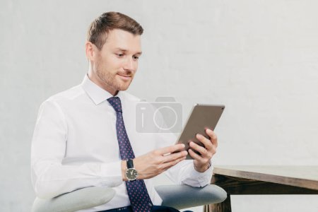 Foto de Gerente ejecutivo masculino en camisa blanca y corbata, sostiene tableta ordenador, busca la información necesaria en Internet, conectado a wifi en la oficina. Concepto de personas, negocios y tecnologías modernas - Imagen libre de derechos