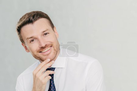 Photo for Horizontal shot of cheerful male with stubble, holds chin, looks positively at camera, dressed in formal shirt with tie, listens something pleasant, isolated over white background. Man employee - Royalty Free Image