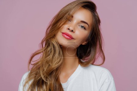 Photo for Portrait of satisfied relaxed young female model tilts head, has makeup, fair hair, dressed in white clothes, poses against purple background, has well cared complexion. People, beauty, face care - Royalty Free Image