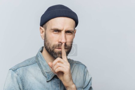Photo for Serious pleasant looking bearded male with mysterious expression keeps fore finger on lips, asks to keep personal information confidential, wears stylish hat and denim shirt, isolated on grey wall - Royalty Free Image