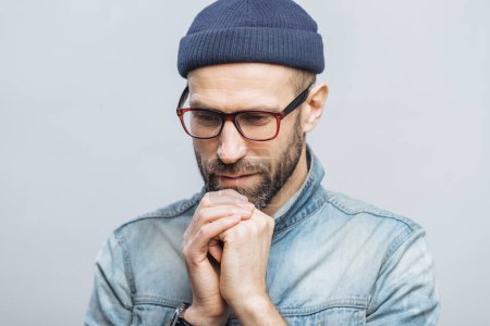 Photo for Shot of hopeful bearded male looks with thoughtful expression down, keeps hands together, hopes for better, worships in calm atmosphere, isolated over white studio background. Horizontal shot - Royalty Free Image
