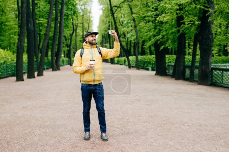 Téléchargez les photos : Portrait horizontal d'un bel homme barbu en anorak jaune, casquette et jean ayant une expression heureuse tout en posant dans un parc faisant du selfie en buvant du café à pied. Hipster étant excité par selfie - en image libre de droit