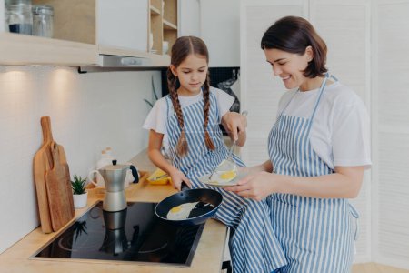 Téléchargez les photos : Photo of cheerful busy mum and her daughter pose near stove, serve breakfast for family, fry eggs on frying pan, wear aprons, pose in domestic kitchen. Mother teaches kid how prepare meal easily - en image libre de droit