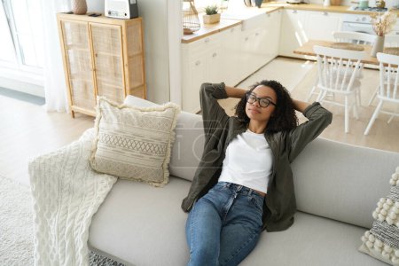 Foto de Calm african american young woman sitting on cozy couch in living room put hands behind head. Relaxed girl resting on comfortable sofa, enjoy fresh conditioned air, lazy weekend at home. - Imagen libre de derechos