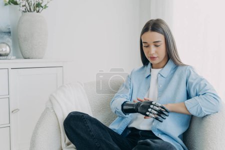 Téléchargez les photos : Handicapped girl looks and touches her bionic arm. Young pensive european woman with cyber hand at home. Modern bionic prosthesis. Futuristic technology of artificial limbs. - en image libre de droit