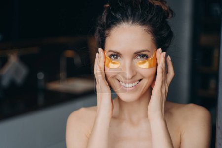 Beautiful hispanic girl doing spa procedures and touches her temples skin. Portrait of happy young lady which applies collagen eye patches. Golden anti age and anti wrinkle patches.