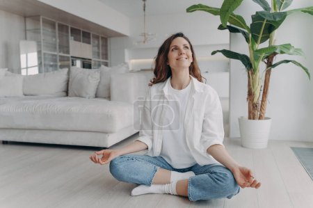 Foto de Peace of mind and mental health concept. Young european woman is practicing yoga on floor and smiling. Meditation, consciousness and relaxation at home. Posture exercise, lotus asana. - Imagen libre de derechos