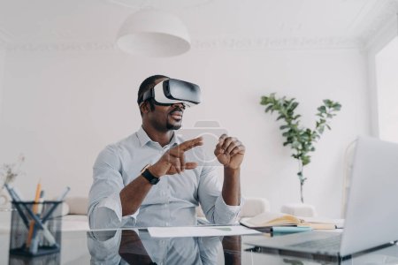 African american businessman using virtual reality glasses sitting at laptop at workplace in office. Modern black guy businessperson wearing VR goggles. Augmented Reality. Future high-tech.