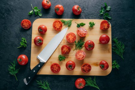 Photo for Top view of red sliced tomatoes on wooden chopping board. Sharp knife near. Green parsley and dill. Dark background. Preparing fresh vegetable salad - Royalty Free Image