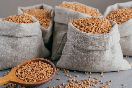 Photo for Close up shot of little sack made of linen fabric filled with dry brown buckwheat, wooden spoon with raw cereal. Selective focus - Royalty Free Image