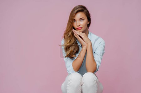 Photo for Serious beautiful woman with long hair, dressed in shirt and trousers, keeps palms pressed together undr chin, has confident calm look, sits on chair over violet background, copy space area aside - Royalty Free Image