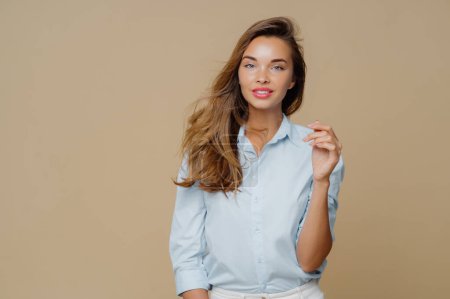 Photo for Pleased lovely woman with long hair, keeps hand raised, wears fashionable shirt, has clam face expression, smiles pleasantly, stands in studio against brown background. People and fashion concept - Royalty Free Image