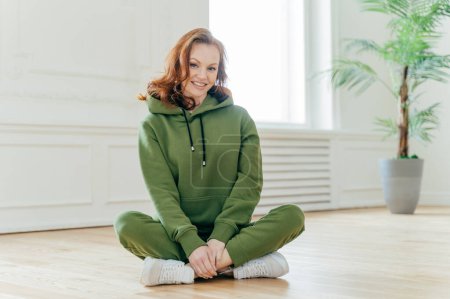 Photo for Happy ginger female sport trainer sits crossed legs on floor, wears green hoody and trousers, white trainers, smiles gladfully, poses on floor in empty room for going in for sport. Healthy lifestyle - Royalty Free Image
