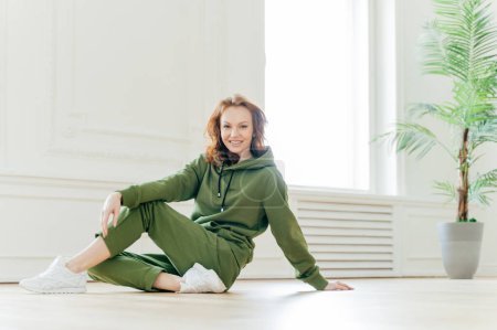 Photo for Happy fitness red haired woman leans with hand on floor, has time for rest after gymnastic exercises, wears green tracksuit, poses indoor, has athletic body. People, sport, lifestyle concept - Royalty Free Image