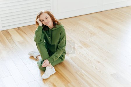 Photo for Pleased restful woman has gentle smile, makeup, wears sportswear, white sneakers, poses on laminate floor, practices yoga or workout exercises, expresses happiness. People and lifestyle concept - Royalty Free Image