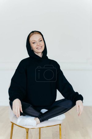 Photo for Relaxed cheerful woman in black sweatshirt, sits in lotus pose on chair, sits indoor, has glad expression, poses indoor, satisfied after active training with coach. People, rest, motivation concept - Royalty Free Image