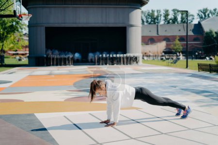 Photo for Full length side view shot of young active woman stands in plank position dressed in sweatshirt leggings sneakers has training outdoors on stadium goes in for sport. Morning workout concept. - Royalty Free Image