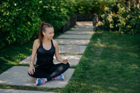 Photo for Athletic sportswoman sits crossed legs on path uses modern smartphone for listening music dressed in active wear chats online poses outdoors during sunny summer day has rest after active training - Royalty Free Image