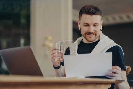 Photo for Man freelancer looking at document in front of laptop, sitting at home office at his work desk, holding glass of water and using earphones, young businessman in casual wear working remotely from home - Royalty Free Image