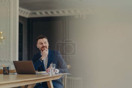 Photo for Successful happy bearded businessman in formal dark blue suit writing down his business plan in notepad while sitting at work desk in front of laptop in stylish light office or luxury apartment - Royalty Free Image