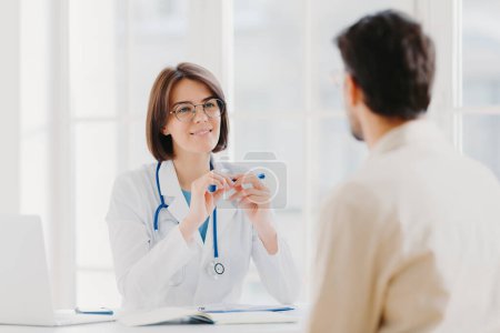 Photo for Professional female doctor in coat listens attentively patients symptoms, examines health, writes down information in patient card, pose in clinic. Sick male tells complaint, sits back to camera - Royalty Free Image