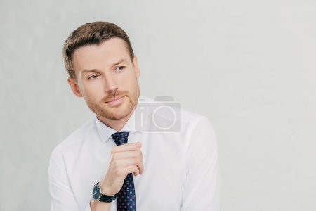 Photo for People and confidence concept. Attractive young unshaven male employee looks thoughtfully aside, contemplates about something important, wears white shirt and tie, isolated over white background - Royalty Free Image