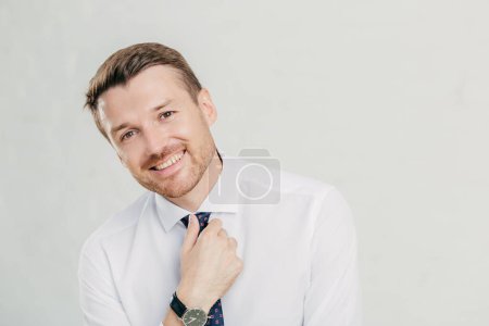 Photo for Cheerful unshaven male keeps hand on tie, dressed in elegant white shirt, smiles friendly, happy to achieve success in life, being prosperous and rich, isolated over white background. Man leader - Royalty Free Image