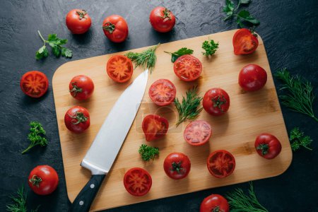 Photo for Tasty ripe tomatoes sliced on chopping board with greenery, sharp knife near, isolated over dark background. Kitchenware. Top view. - Royalty Free Image