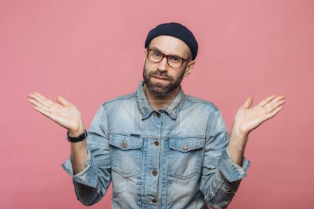 Photo for Portrait of hesitant bearded male with doubtful expression, shrugs shoulders, wears denim jacket, glasses and hat, isolated over pink studio background. Confused middle aged man poses indoor - Royalty Free Image