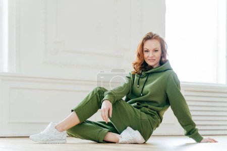 Photo for Sideways shot of satisfied redhead woman has makeup, wears tracksuit, white spotshoes, rests on floor, does stretching exercises, is fond of sport. People, flexibility and motivation concept - Royalty Free Image