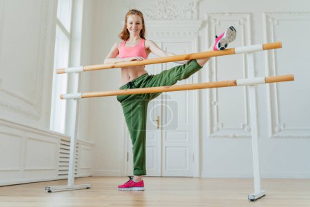 Photo for Indoor shot of redhead woman dancer stretches legs on barre in ballet studio, wears pink top and trousers, pink sneakers, does pilates fitness exercises, poses in gym studio, prepares muscles - Royalty Free Image
