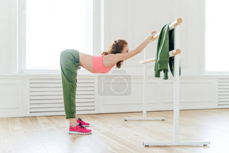 Photo for Indoor shot of sportswoman in sportsbra and active wear, leans at ballet barre, has productive training session, slim body shape, stretches muscles, has motivation for being fit and healthy. - Royalty Free Image