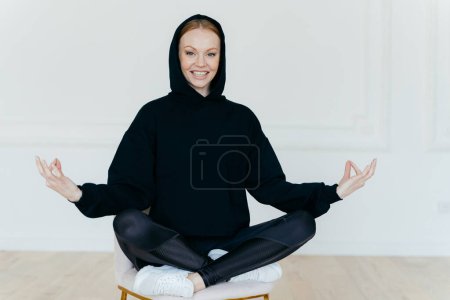 Photo for Horizontal shot of optimistic woman practices yoga, meditates on chair, sits in lotus pose, wears black sweatshirt, white sportshoes, has glad expression, tries to relax after hard working day - Royalty Free Image