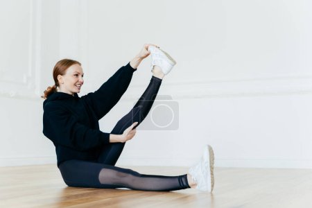 Photo for Cheerful fit woman raises leg, stretches before fitness and exercise, has athletic body, wears black sportcostume, sneakers, sits on floor, has great progress, has ginger hair with pony tail. - Royalty Free Image