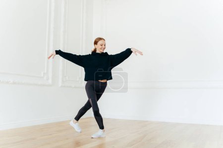 Photo for Professional female ballerina practices dance in hall, stretches hands, stands with crossed legs, has gentle smile on face, dressed in black sportswear, enjoys spare time for hobby. Healthy lifestyle - Royalty Free Image