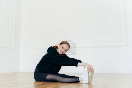 Photo for Delighted young athletic girl does stretching exercises, prepares muscles before training, dressed in black hoody and comforatble leggings, sits on floor. People, workout, sporty life concept - Royalty Free Image