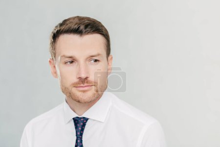 Photo for Pensive young male businessman looks thoughtfully aside, thinks about business project, focused into distance, isolated over white background with copy space aside for advertisement or text. - Royalty Free Image