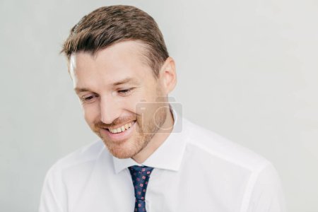 Photo for Joyful male corporate worker in elegant white shirt, looks happily down, remembers something pleasant in mind, has toothy smile, wears elegant white shirt with tie, isolated over white wall. - Royalty Free Image