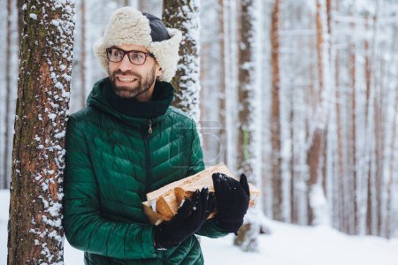 Téléchargez les photos : Le jeune homme barbu porte un chapeau chaud et une veste verte, tient une pile de bois de chauffage, regarde joyeusement de côté, remarque quelque chose, aime l'hiver et le temps glacial. Homme attrayant rêveur passe du temps dans la forêt - en image libre de droit