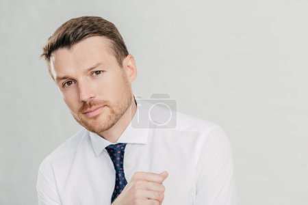 Photo for Horizontal shot of handsome young male entrepreneur in formal white shirt, looks directly at camera, listens attentively his interlocutor, talk on business theme, isolated over white background - Royalty Free Image