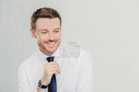 Photo for Professional male office manager giggles positively, concentrated down, shows white teeth, dressed in white shirt and tie, remembers something pleasant, isolated over white background with copy space - Royalty Free Image