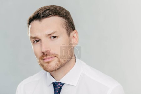 Photo for Close up portrait of handsome unshaven man looks seriously at camera, has appealing look, thinks about business affairs, dressed in formal clothes, isolated over white background. CEO indoor - Royalty Free Image