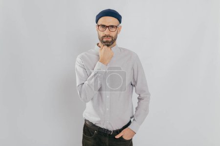 Photo for Waist up shot of serious man keeps one hand under chin, other in pocket, has stubble, wears hat, white shirt, trousers, isolated over white background, listens something attentively. Masculinity - Royalty Free Image