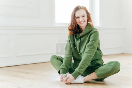 Photo for Indoor shot of beautiful redhead European woman has rest after cardio training, keeps legs crossed, dressed in green tracksuit, sits on floor alone. Healthy lifestyle, youth and fitness concept. - Royalty Free Image