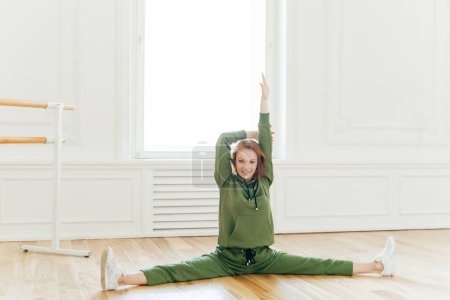 Photo for Full length shot of cheerful redhead woman raises hands, does splits, stretches muscles, sits on floor, stretches legs sideways, wears green tracksuit and sportshoes. Sport and active llifestyle - Royalty Free Image