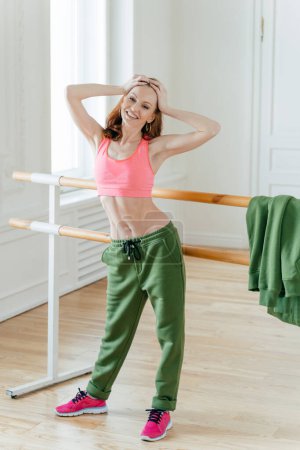 Photo for Red haired woman has perfect figure, keeps hands on head, feels tired after dance rehearsal, stands near ballet barre, wears top, sport trousers and sneakers, poses in dancing studio has smile on face - Royalty Free Image