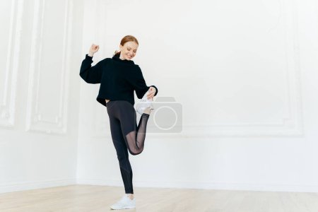 Photo for Full length shot of satisfied slim young woman has makeup stands on one leg, raises feet, dressed in black sportclothes, has fit healthy body shape, glad expression, demosntrates nice flaxibility. - Royalty Free Image