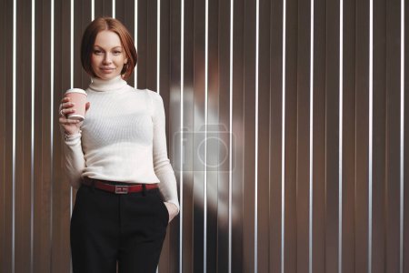 Photo for Half length shot of pleasant looking businesswoman dressed in formal clothes, keeps hand in pocket, drinks takeaway coffee, stands against brown wall with free space on left for your advertisement - Royalty Free Image