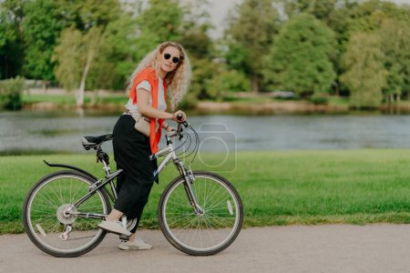 Photo for Horizontal shot of active female bicyclist wears t shirt, black pants and sneakers, stops near river side, keeps feet on pedals of bicycle beautiful scenic nature in background. Sunny day, active rest - Royalty Free Image