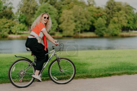 Photo for Outdoor shot of happy blonde curly woman wears sunglasses, dressed in casual active wears, rides on bicycle near lake and green trees in countryside, spends free time outside, enjoys favourite hobby - Royalty Free Image